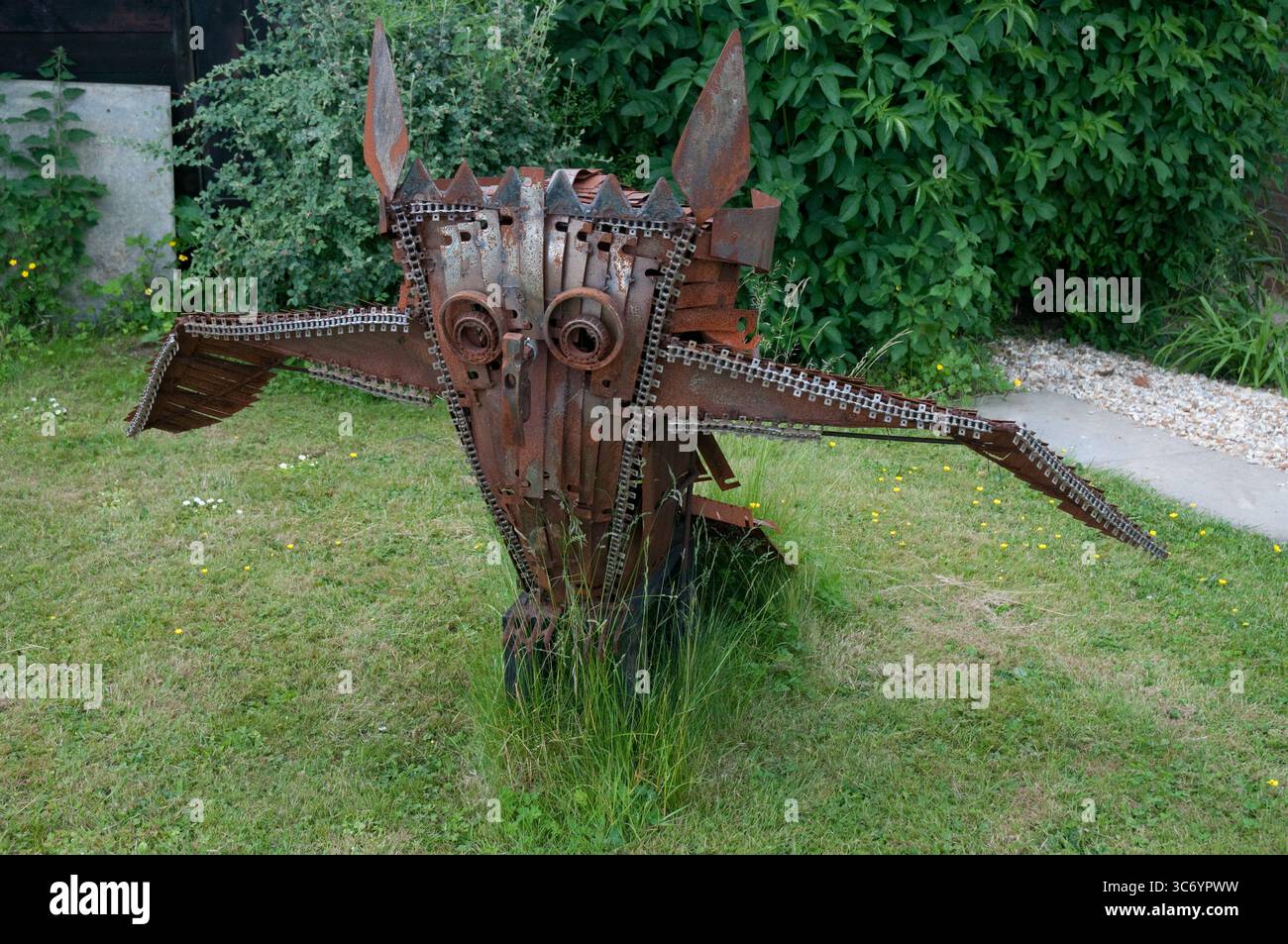 Una grande scultura in metallo simile a un gufo è posizionata in un giardino, completata da piante vibranti e erba sotto la luce del sole. Foto Stock
