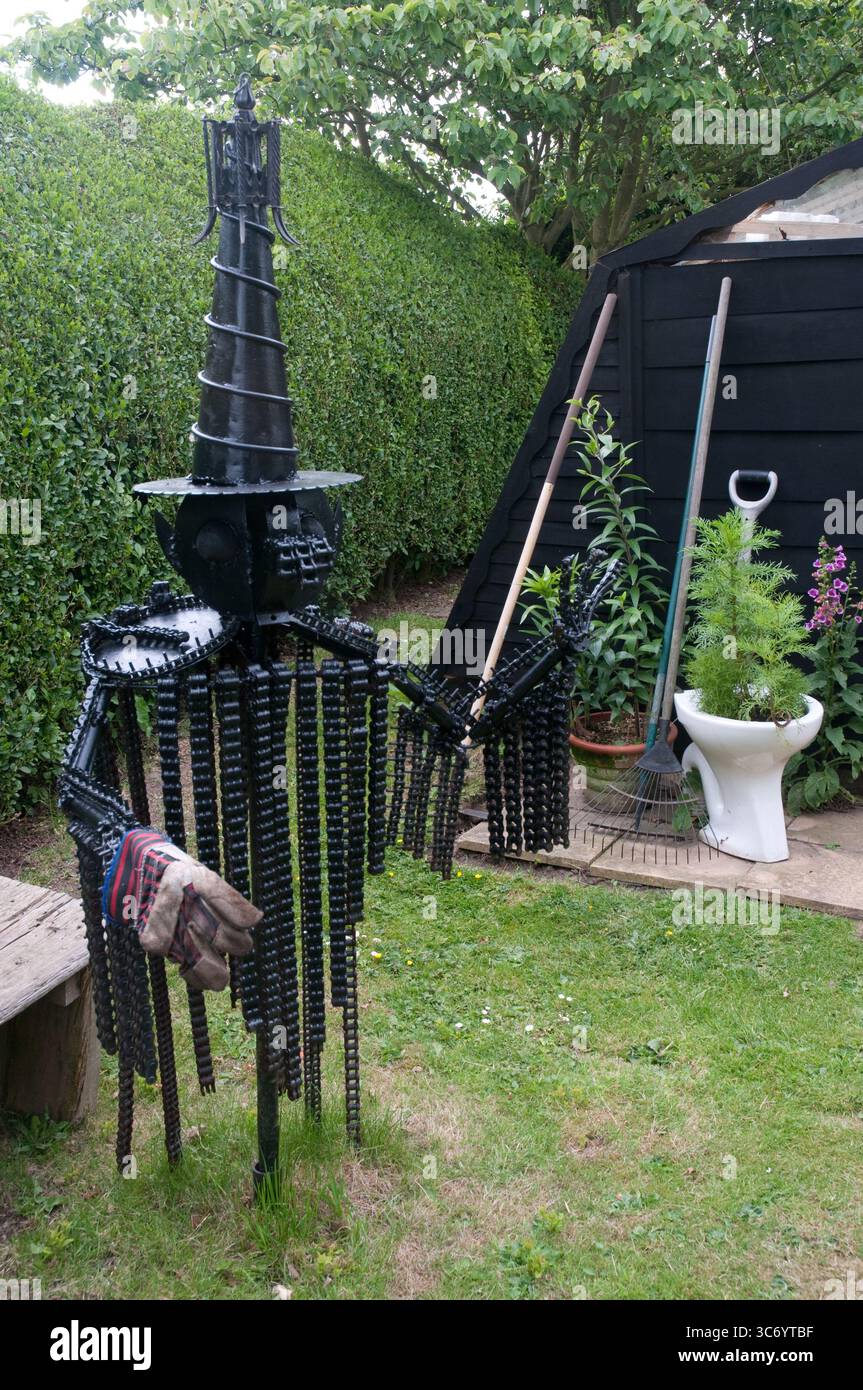 Una scultura in metallo unica che ricorda una figura spettrale si erge in un giardino circondato da verde e strumenti da giardinaggio, mostrando creatività artistica. Foto Stock