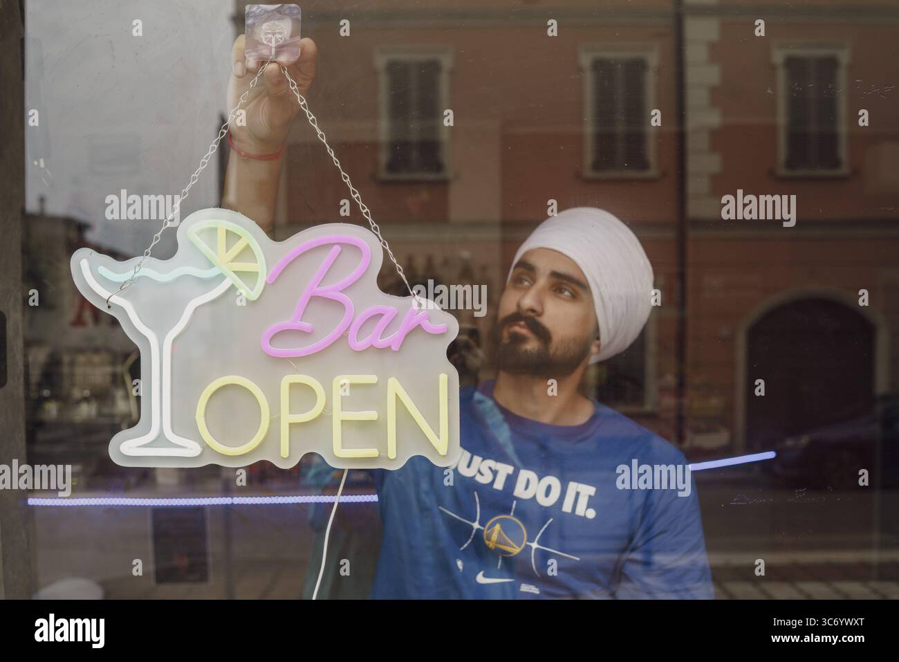 Una persona installa un cartello al neon "Bar Open" su una finestra che riflette un ambiente urbano, Cremona, Italia 25 luglio 2025 Foto Stock
