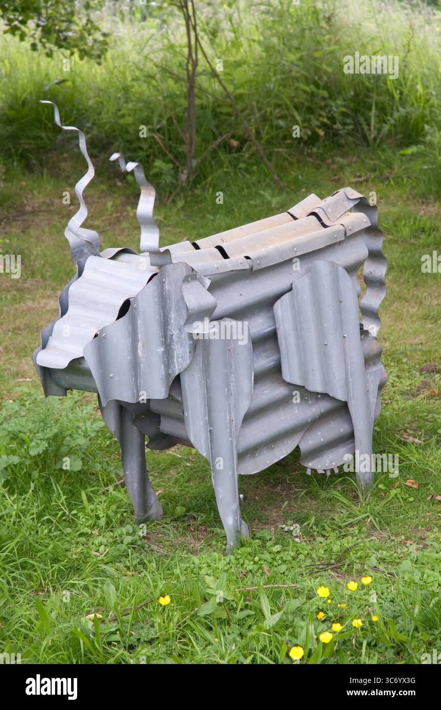 Un'opera d'arte distintiva che assomiglia a una mucca realizzata in metallo ondulato si trova in mezzo all'erba e ai fiori selvatici in un giardino. Foto Stock