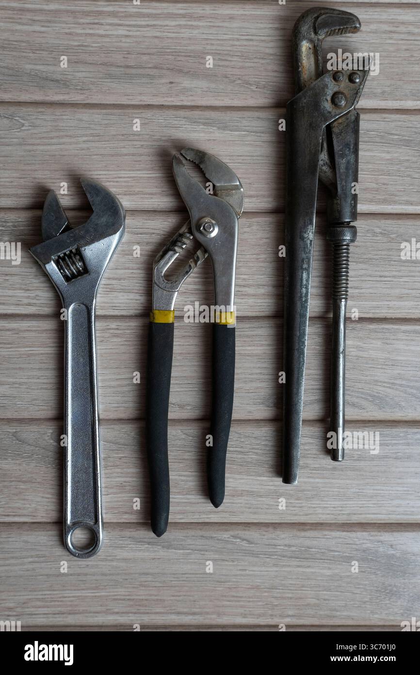 Disposizione piatta ad alta risoluzione di tre utensili manuali in metallo (chiave regolabile, pinza per giunti scorrevoli e chiave per tubi) disposti su una superficie rustica in legno Foto Stock