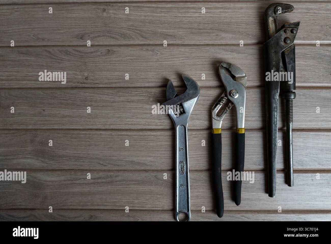 Tre utensili manuali in metallo, una chiave per tubi, una chiave regolabile e una pinza per giunti scorrevoli, disposti su uno sfondo in legno resistente agli agenti atmosferici Foto Stock