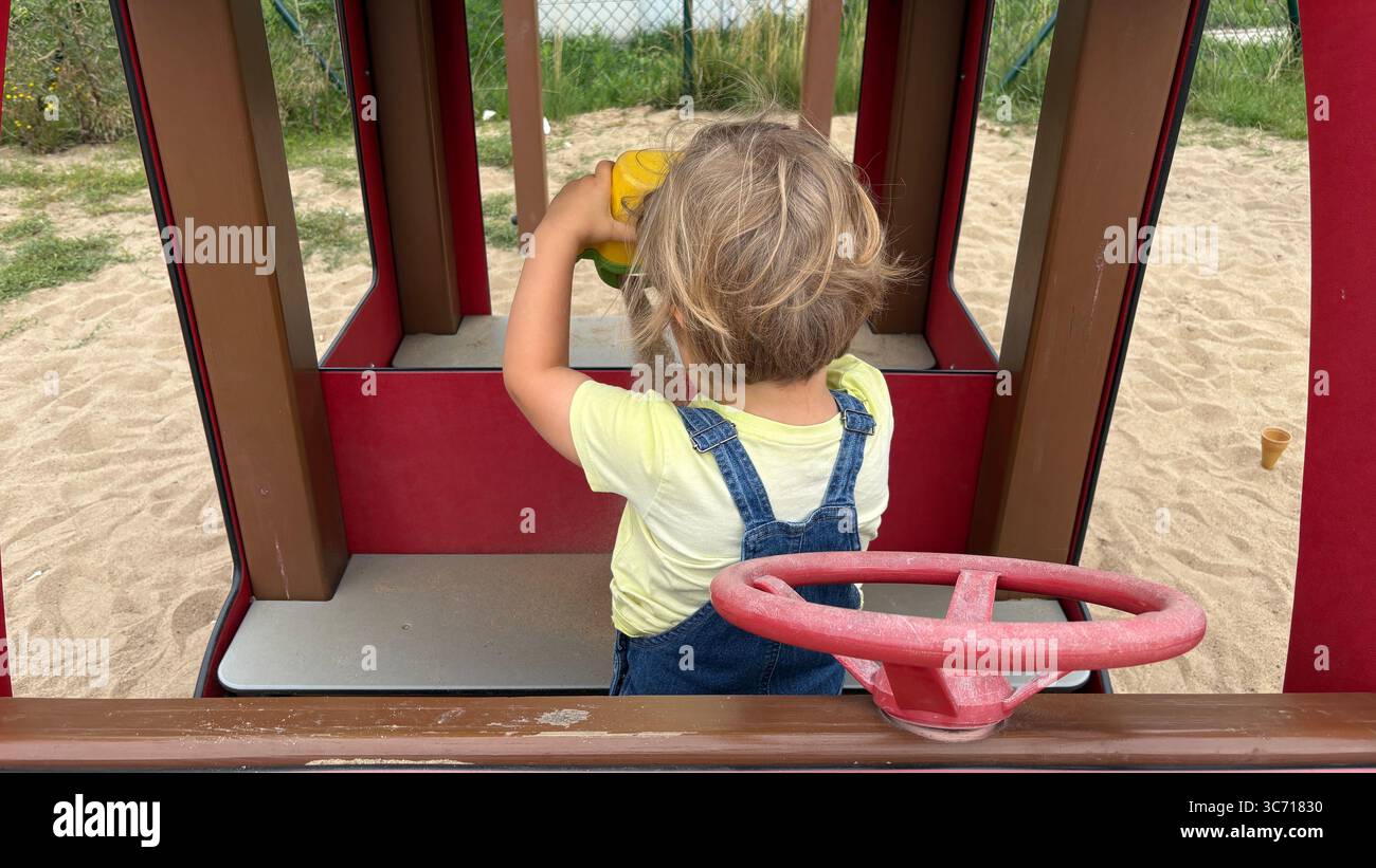 Il bambino versa sabbia dalla tazza giocattolo all'interno di un parco giochi, impegnandosi in un gioco all'aperto fantasioso e nell'esplorazione sensoriale. Foto Stock