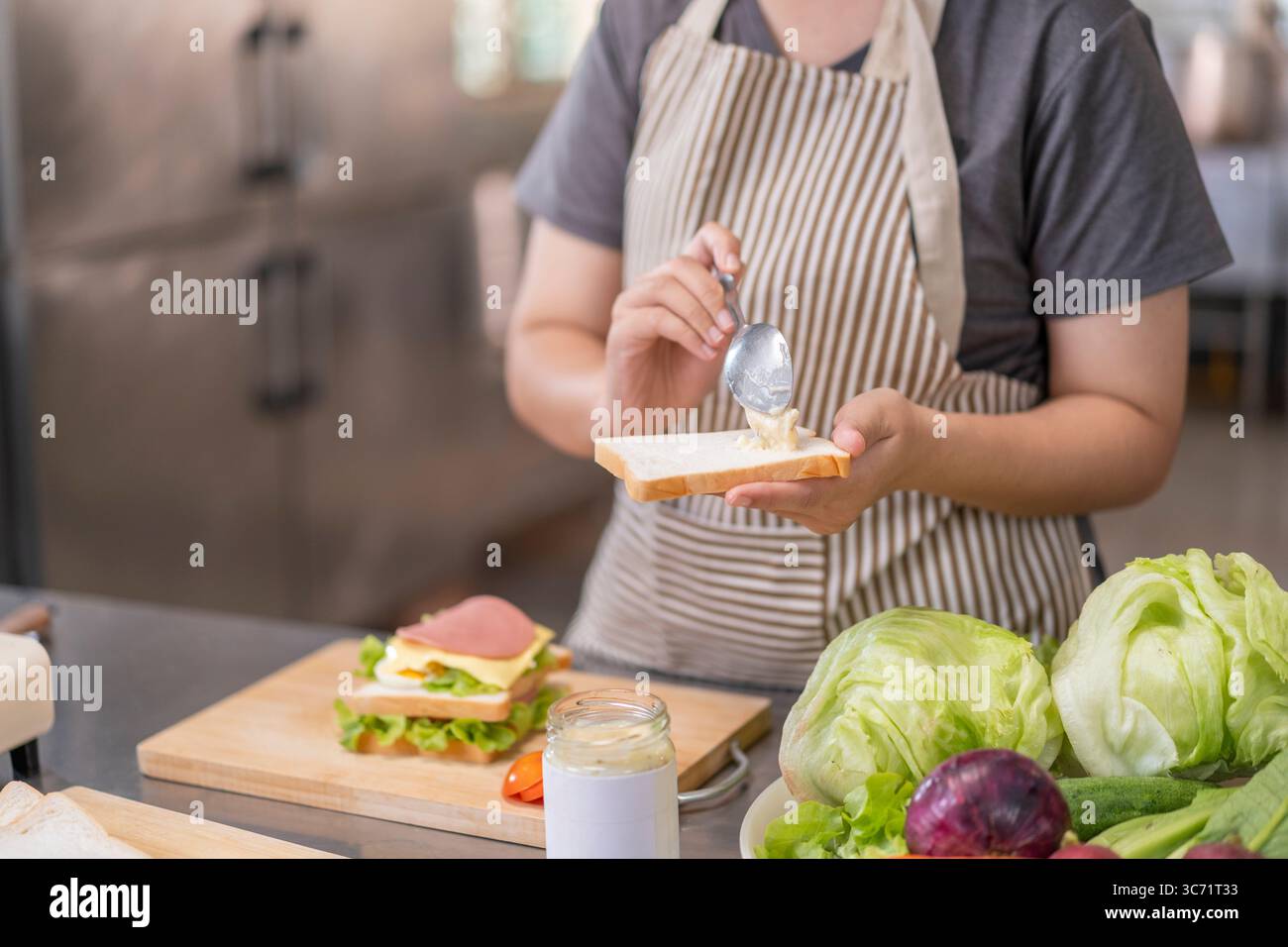 Una giovane donna asiatica, che ha 20 anni, indossa un grembiule a righe, usa un cucchiaio per spalmare la maionese su una fetta di pane mentre prepara una casa deliziosa e sana Foto Stock