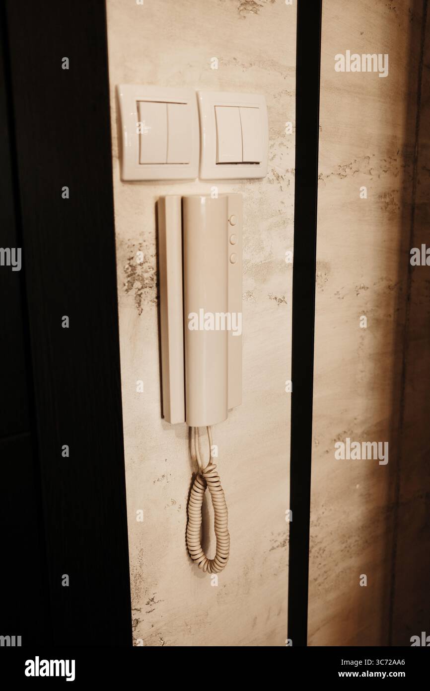 Telefono interfono montato a parete con cavo a spirale accanto a due interruttori luminosi su una parete testurizzata nel corridoio. Foto Stock