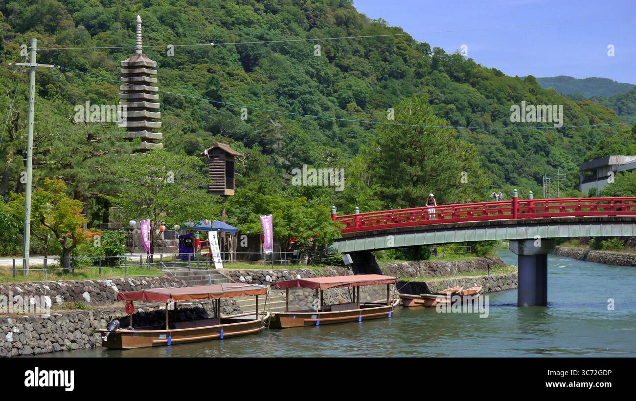 Vista panoramica del fiume Uji in Giappone con barche tradizionali, ponte rosso e torre di pagoda circondata da lussureggianti montagne verdi. Foto Stock