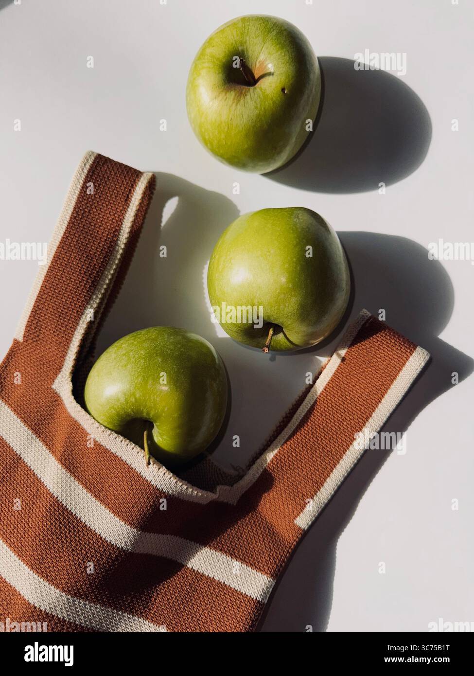 Tre mele verdi e una borsa in cotone a righe su superficie bianca con ombre morbide e un'estetica minimalista alla luce del sole naturale Foto Stock