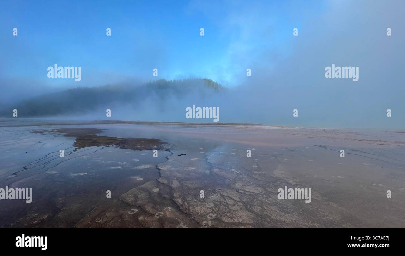 Un paesaggio sereno presenta una sorgente termale con la nebbia che si avvolge sopra di esso e uno sfondo di colline boscose sotto un cielo blu brillante. Foto Stock