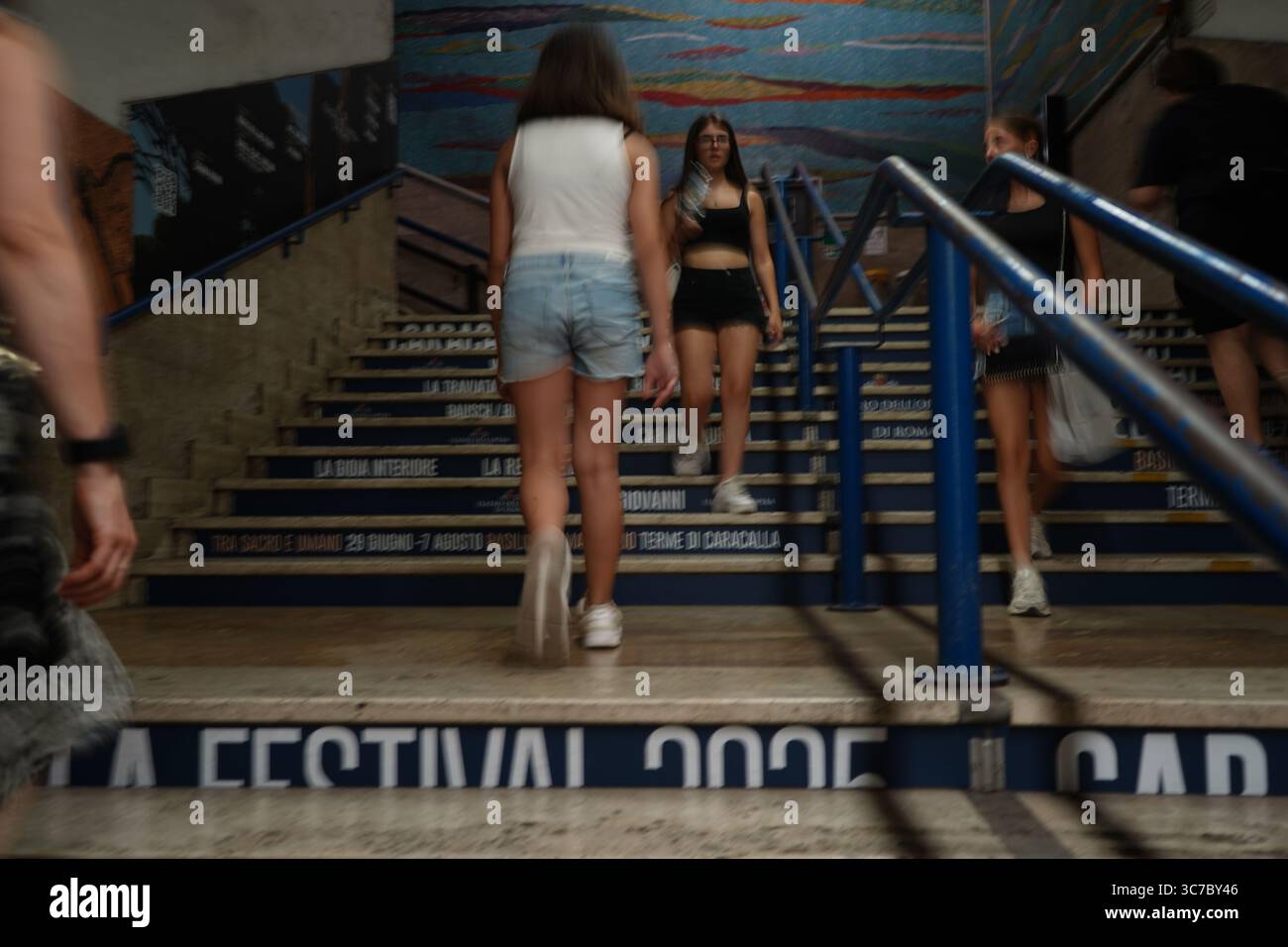 Roma, Italia, 13 luglio 2025. Persone sfocate che camminano su una scala con sfondo a mosaico in una stazione della metropolitana di Roma. Foto Stock