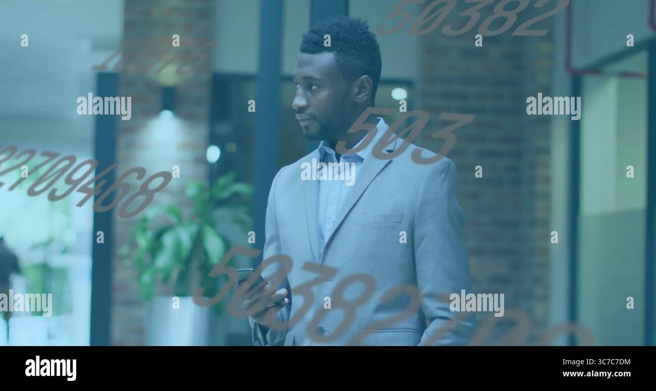 Uomo in tuta grigio chiaro che tiene in mano lo smartphone mentre guarda a sinistra nel corridoio dell'ufficio, con piante in vaso Foto Stock