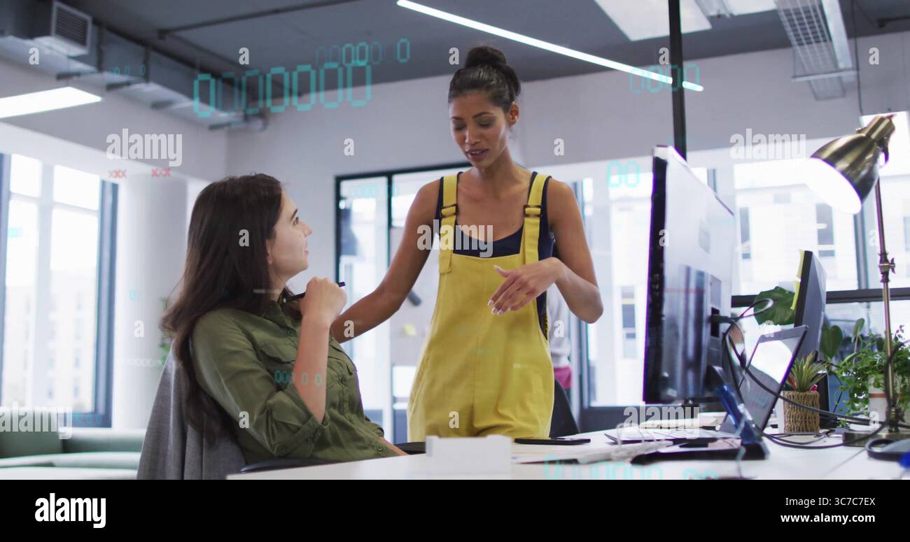 Parlare con un collega in piedi e fare gesto verso un collega seduto in un ufficio open space, con due monitor Foto Stock