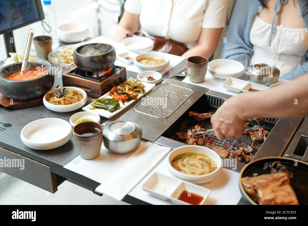 In un ristorante barbecue kalbi, un server grigia un pezzo di carne su carboni scintillanti, fiamme che leccano i bordi mentre il vapore sale dal grasso frizzante, riempiendo la A. Foto Stock