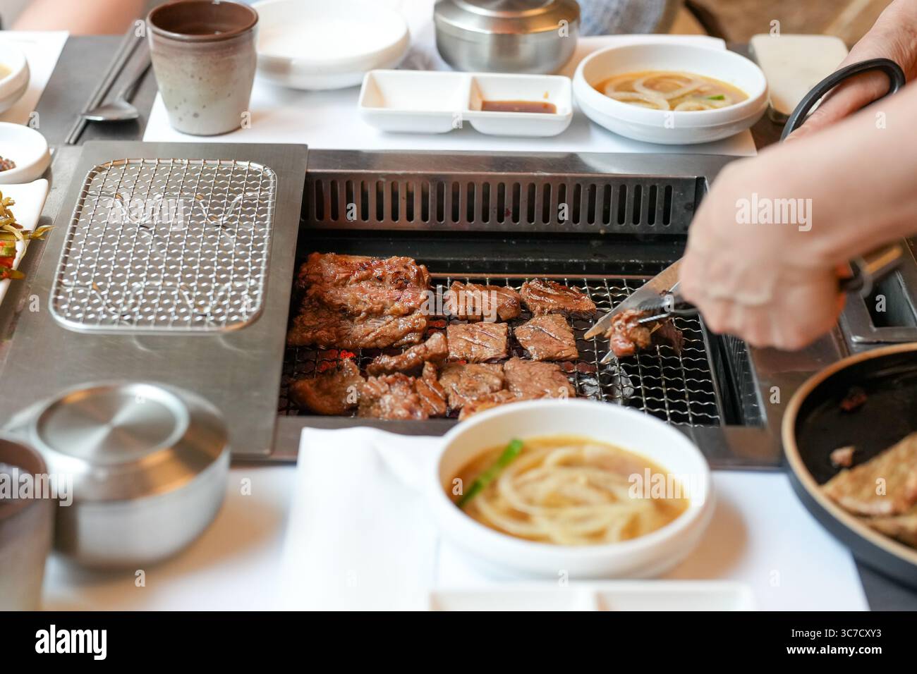 In un ristorante barbecue kalbi, un server grigia un pezzo di carne su carboni scintillanti, fiamme che leccano i bordi mentre il vapore sale dal grasso frizzante, riempiendo la A. Foto Stock