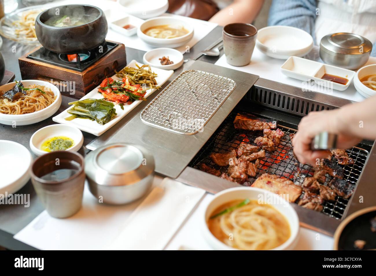 In un ristorante barbecue kalbi, un server grigia un pezzo di carne su carboni scintillanti, fiamme che leccano i bordi mentre il vapore sale dal grasso frizzante, riempiendo la A. Foto Stock