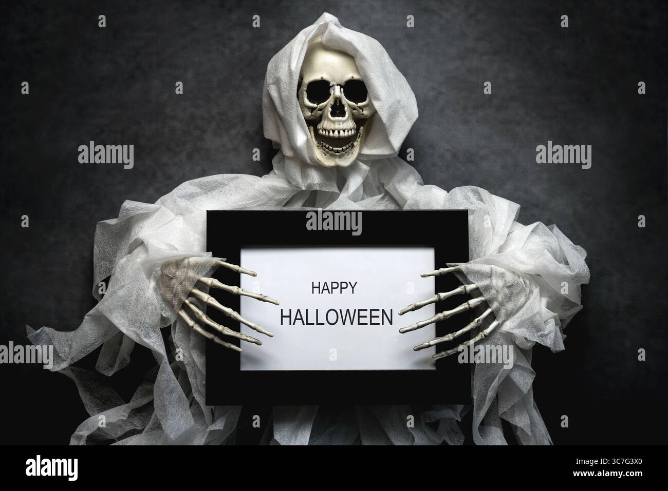 Spaventoso scheletro malvagio sorridente che regge una cornice nera con il testo Happy Halloween su sfondo scuro. Sfondo del concetto di Halloween Foto Stock