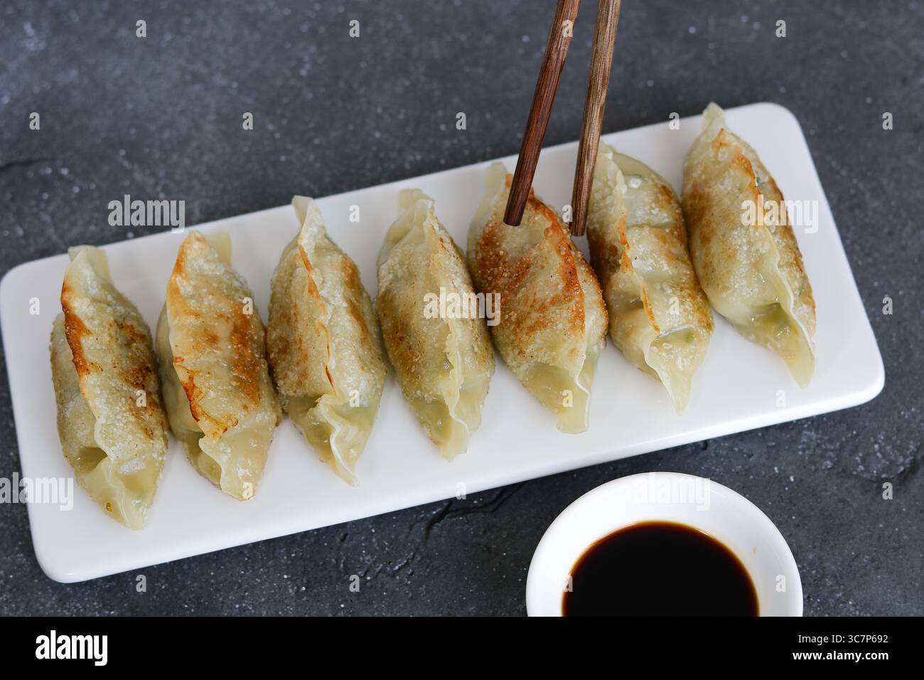 Mano che tiene un paio di bacchette per mangiare gnocchi Mandu o coreani. Accanto al piatto è presente un piccolo recipiente di salsa di soia scura. Foto Stock