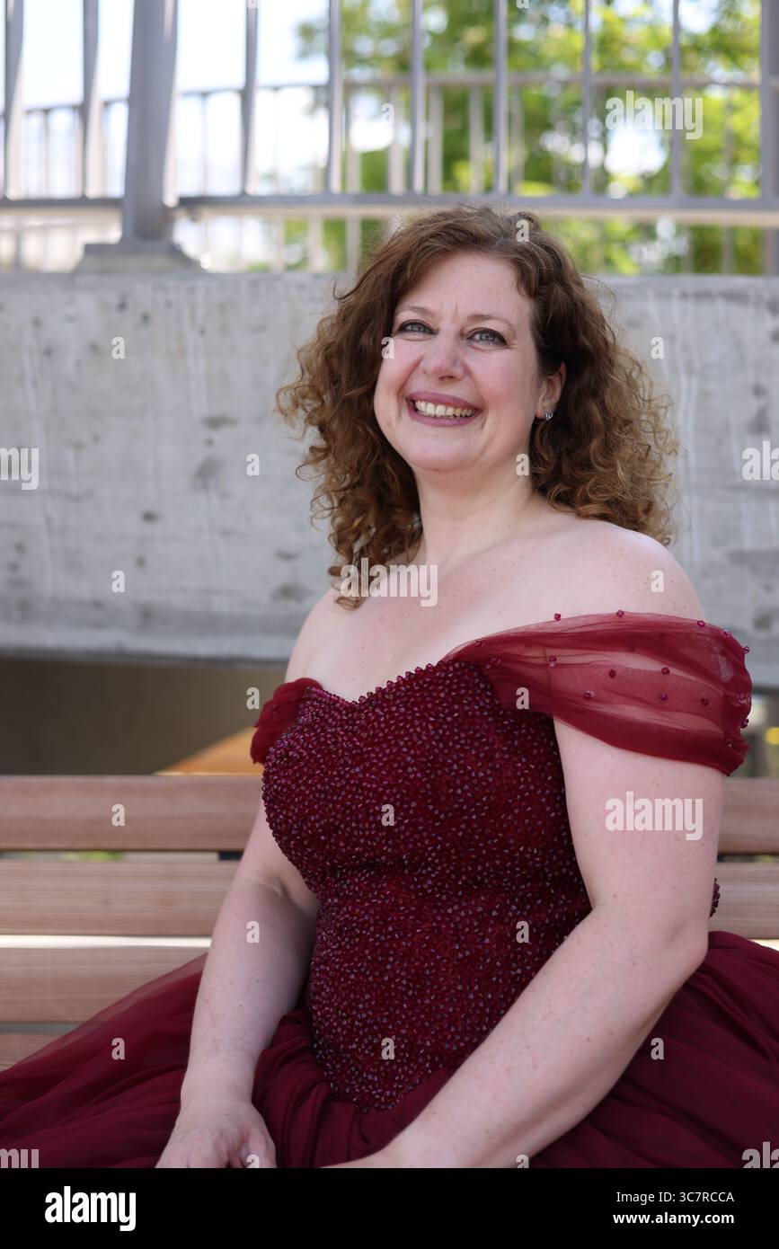 Donna con un pomposo abito da sera rosso seduto sulla panchina e sorride Foto Stock