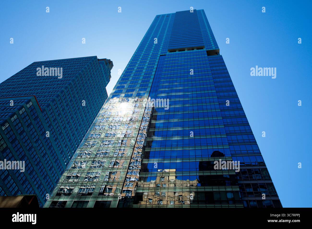 Una torre di vetro a specchio nello skyline di Midtown di Manhattan riflette i vicini edifici illuminati dal sole sotto un cielo azzurro, evidenziando i dinamici contrasti urbani. Foto Stock