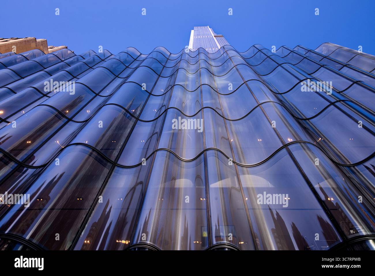 La cortina di vetro ricurva di un edificio di Midtown Manhattan ondeggia verso il cielo, riflettendo delicatamente i dettagli a livello della strada su un vibrante sfondo blu. Foto Stock