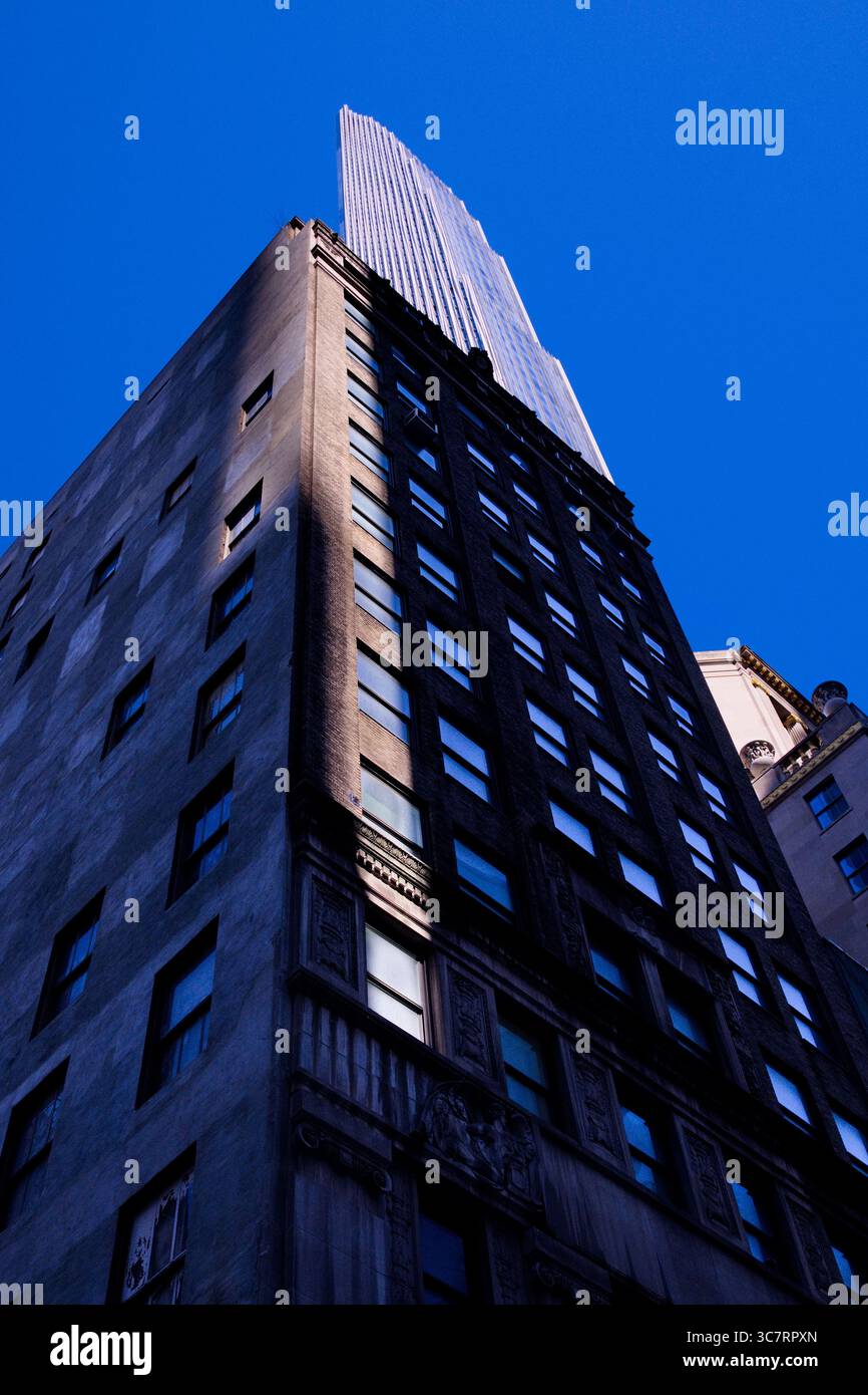Una torre in muratura scura nel canyon di Manhattan si innalza oltre le finestre d'epoca verso una guglia di vetro che mostra facciate testurizzate contro un cielo azzurro. Foto Stock