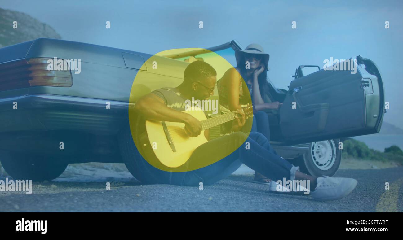 Uomo che suona la chitarra acustica sulla strada costiera vicino alla cabriolet argentata, con cappello e occhiali da sole Foto Stock