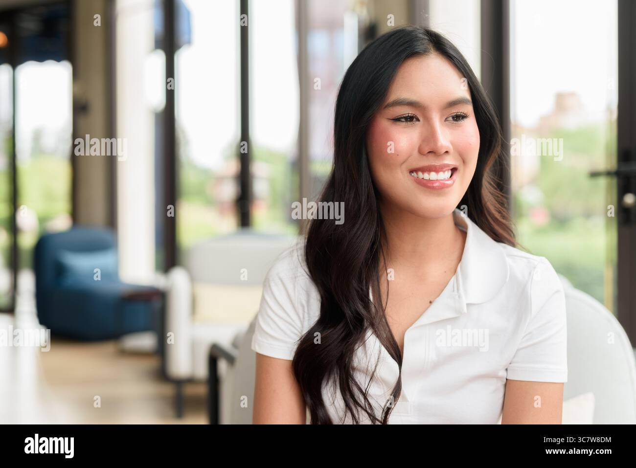 Bella giovane donna asiatica che indossa una polo bianca in posa all'interno dell'hotel o della sala ristorante. Ritratto di stile di viaggio rilassato con interni moderni Foto Stock