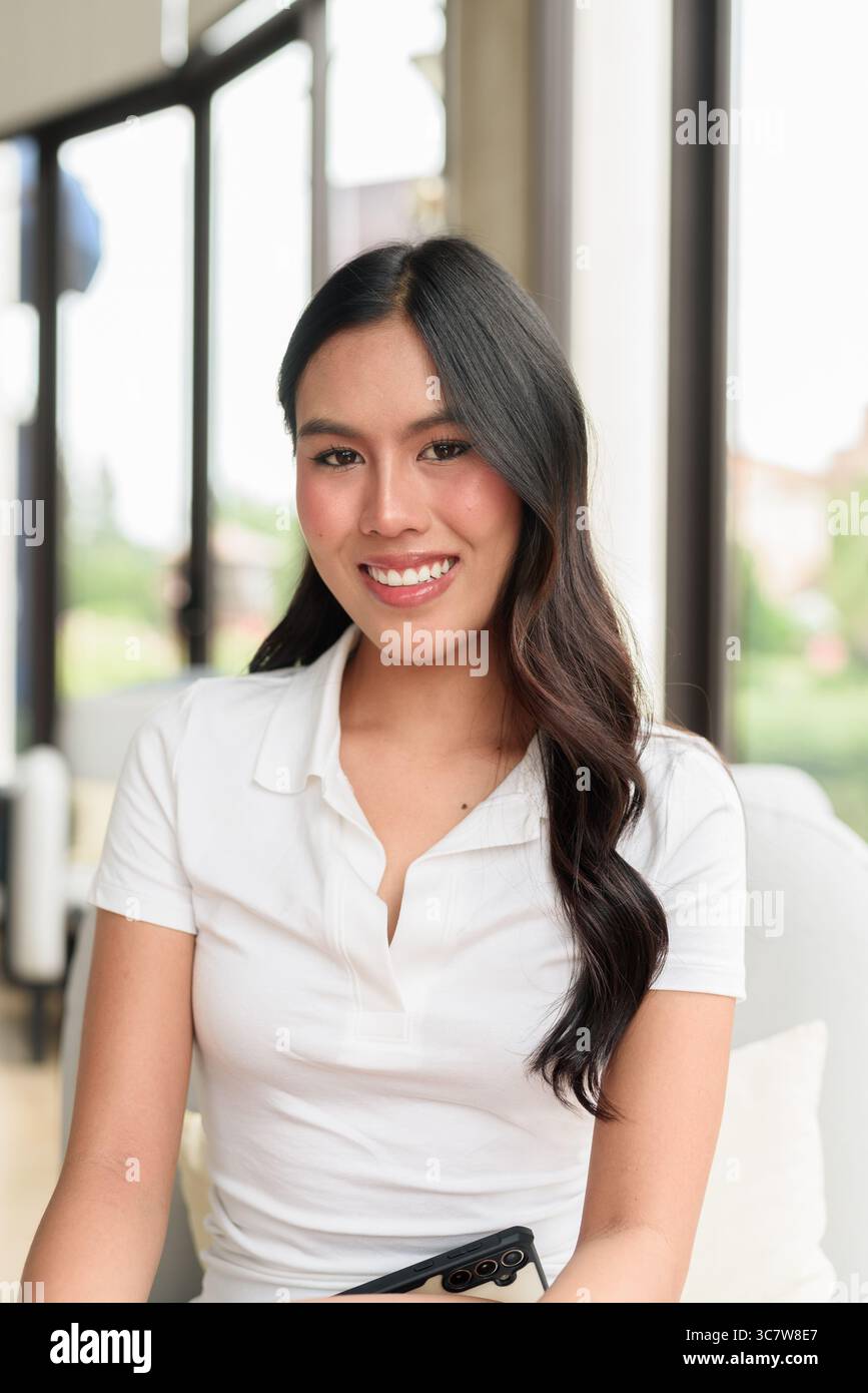 Bella giovane donna asiatica che indossa una polo bianca in posa all'interno dell'hotel o della sala ristorante. Ritratto di stile di viaggio rilassato con interni moderni Foto Stock