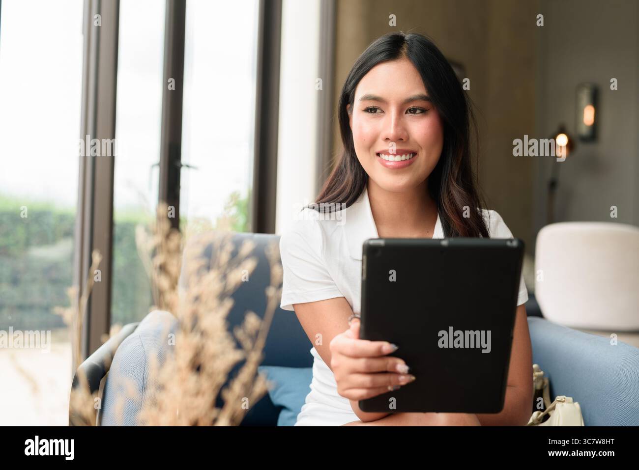 Un giovane e bellissimo turista asiatico che usa un tablet mentre si siede nell'hotel o nella caffetteria lounge. Stile di vita digitale informale ed elegante con interni moderni Foto Stock