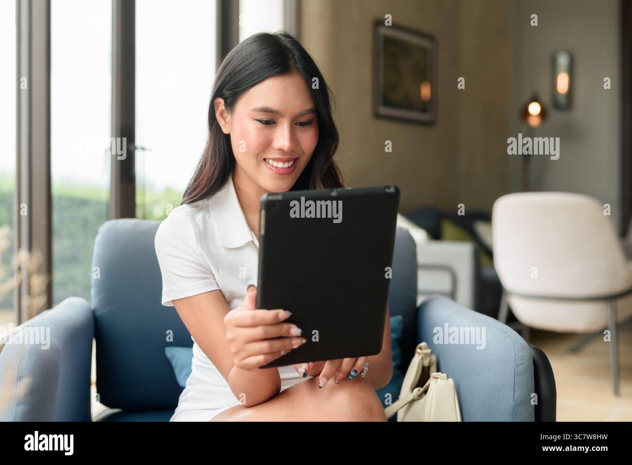 Un giovane e bellissimo turista asiatico che usa un tablet mentre si siede nell'hotel o nella caffetteria lounge. Stile di vita digitale informale ed elegante con interni moderni Foto Stock