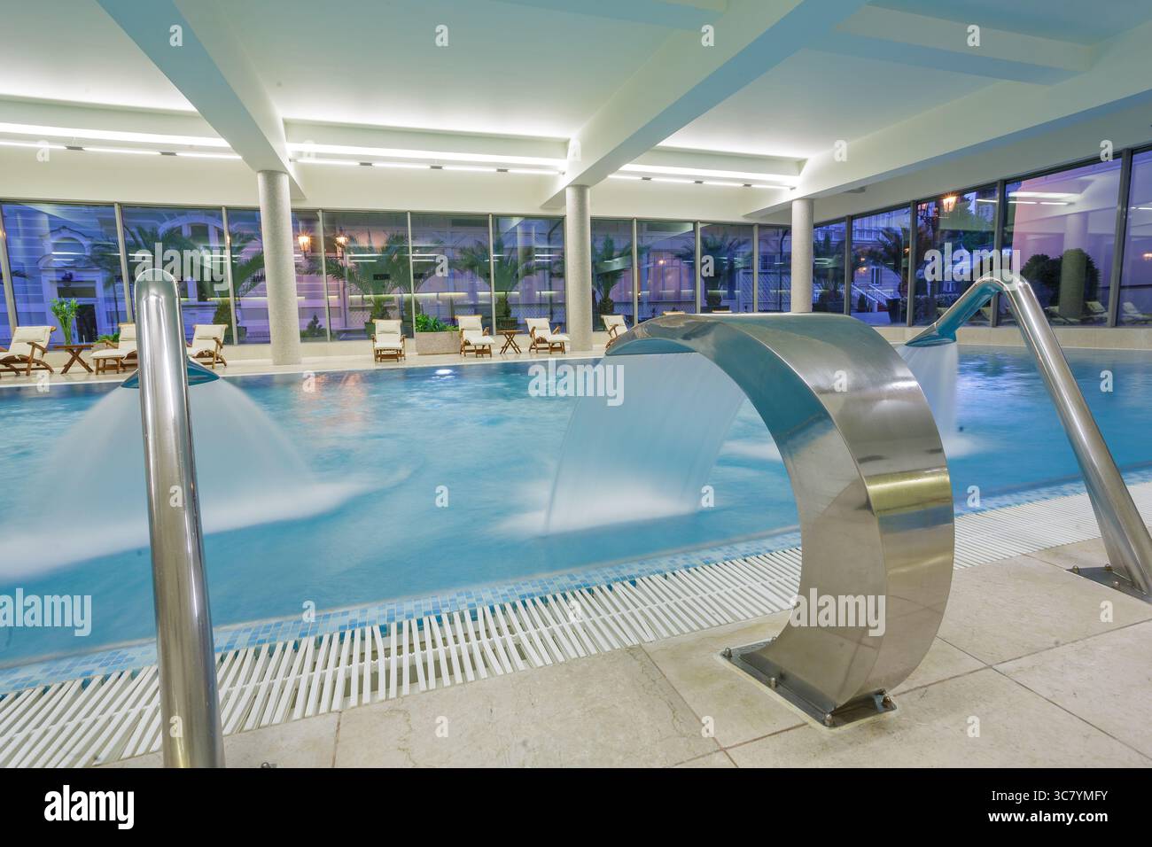 Ampia piscina con idromassaggio al coperto con cascata e rilassanti poltrone nel moderno e luminoso centro benessere dell'hotel Foto Stock