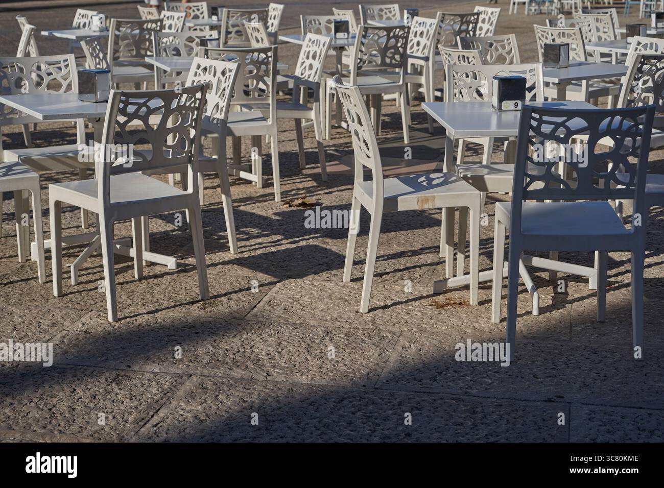 File di sedie decorative contemporanee con motivi geometrici disposte ai tavoli da caffe' sulla piazza di pietra sotto la calda luce del sole Foto Stock