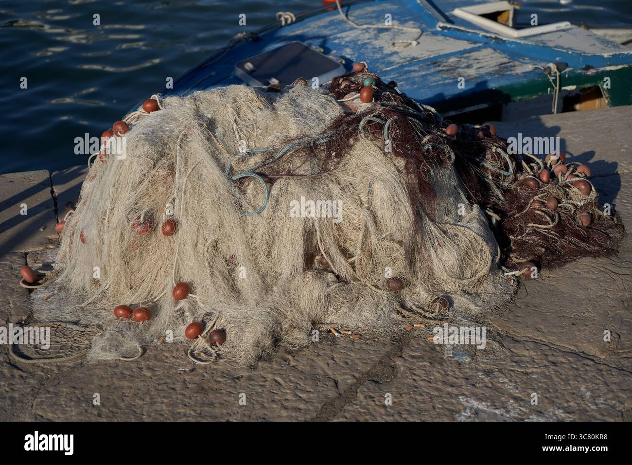 Un mucchio di reti da pesca aggrovigliate con galleggianti in sughero arancione che si asciugano su banchine di pietra accanto al peschereccio blu d'epoca nel porto costiero Foto Stock