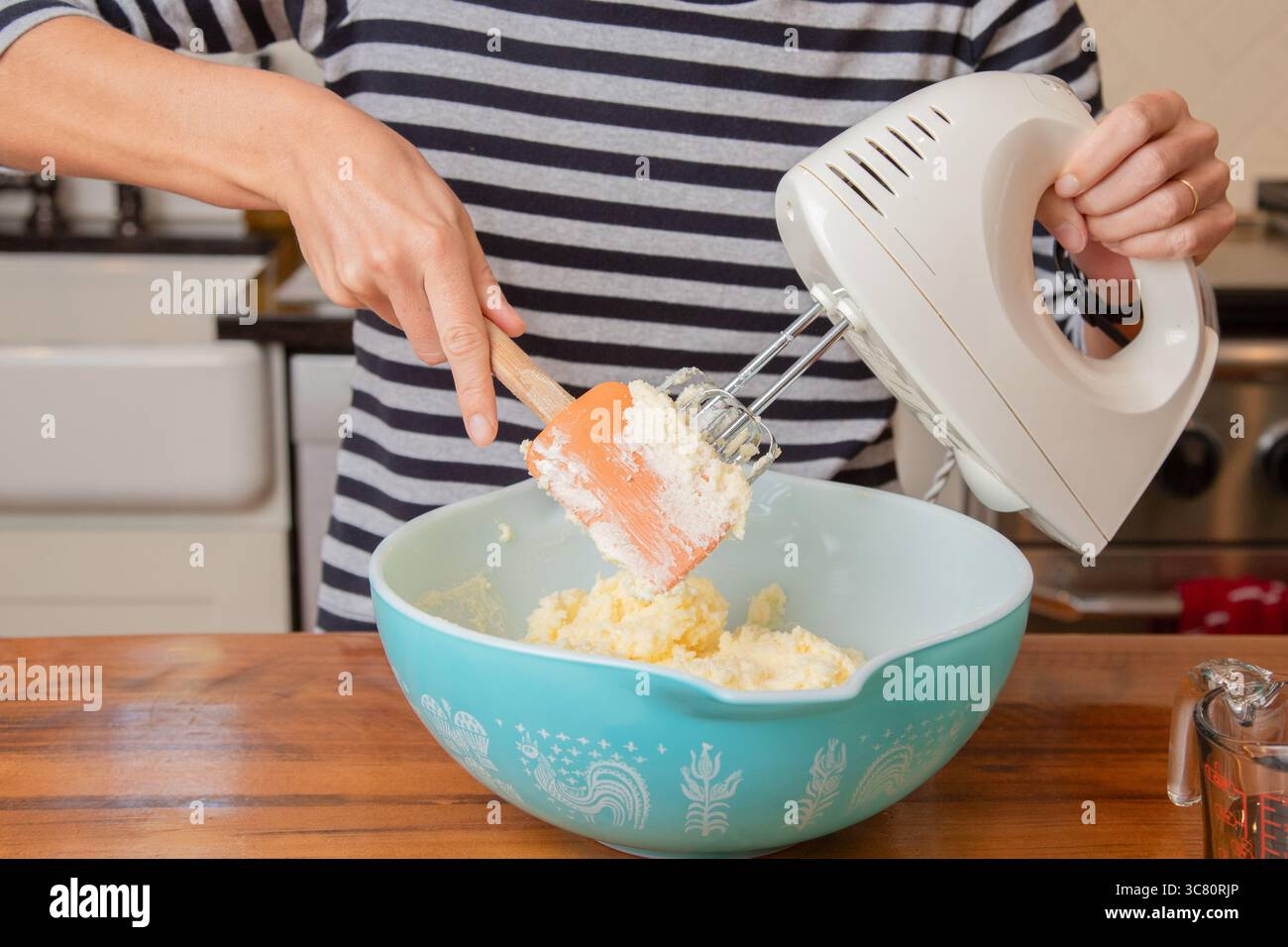 Primo piano di una donna adulta in una cucina che mescola l'impasto in un recipiente con una frusta elettrica e una spatola Foto Stock