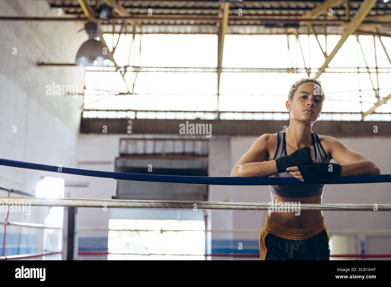 Atleta che si appoggia sulle corde da boxe sotto la luce naturale in palestra con impacchi a mano e spazio per copiare Foto Stock