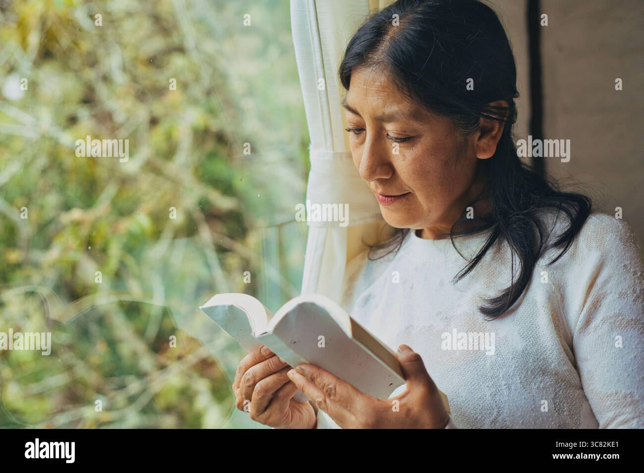 Donna di mezza età che si concentra mentre legge un libro, in piedi accanto a una finestra con luce naturale che illumina la scena, creando un'atmosfera accogliente Foto Stock