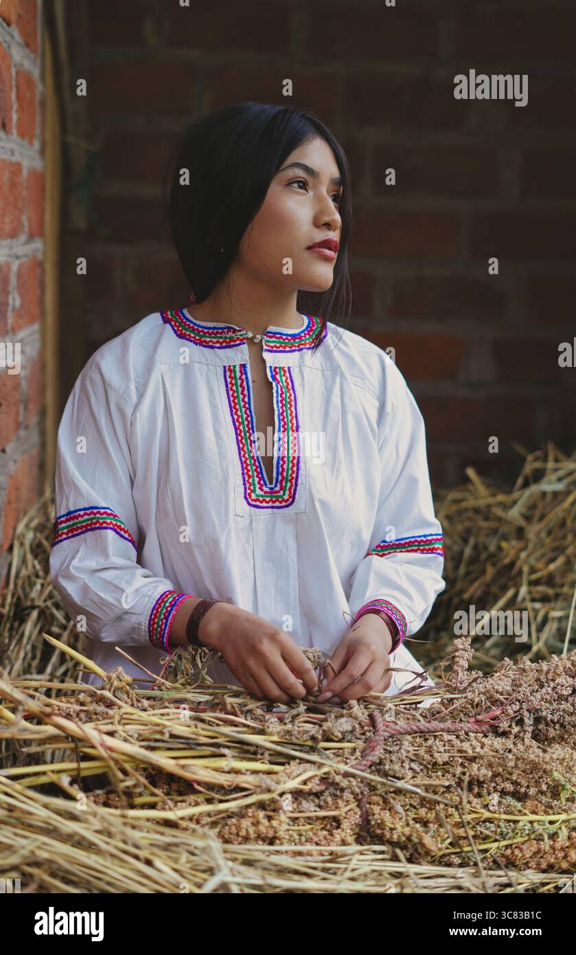 Giovane donna adulta in abbigliamento tradizionale seduta con cura in uno spazio rustico, circondata da pareti di paglia e mattoni, che riflettono il patrimonio culturale e. Foto Stock