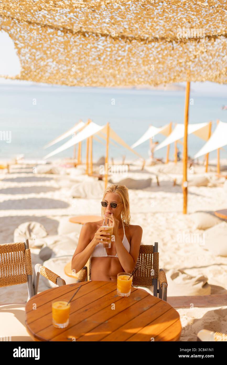 Rilassandosi su una spiaggia soleggiata, una donna sorseggia un drink rinfrescante seduto a un tavolo di legno. I baldacchini bianchi forniscono ombra, creando un perfetto g tropicale Foto Stock