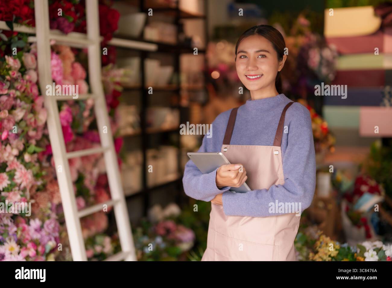 Donna asiatica adulta fiorista gestisce il suo negozio di fiori. Utilizza un tablet per le operazioni aziendali. La sua allegra presenza offre fiori freschi e accogliente Foto Stock