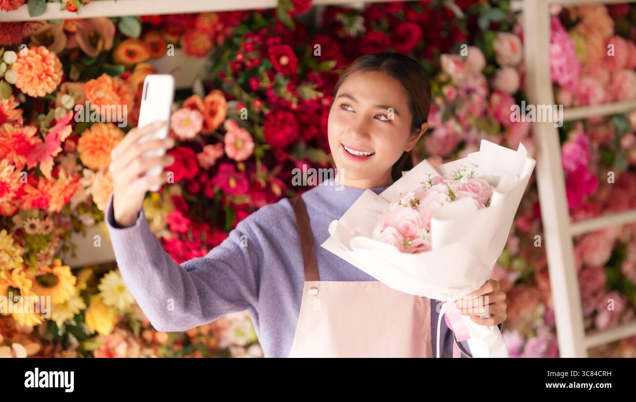 Allegro fiorista adulto asiatico cattura una piacevole composizione floreale con il telefono in un vivace negozio che coinvolge i clienti che promuovono fiori e prodotti Foto Stock