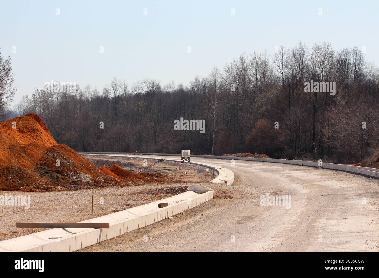Una strada di ghiaia tortuosa con cordoli appena installati si snoda verso un bordo della foresta, mostrando opere di terra attive, attrezzature e materie prime per le zone rurali Foto Stock