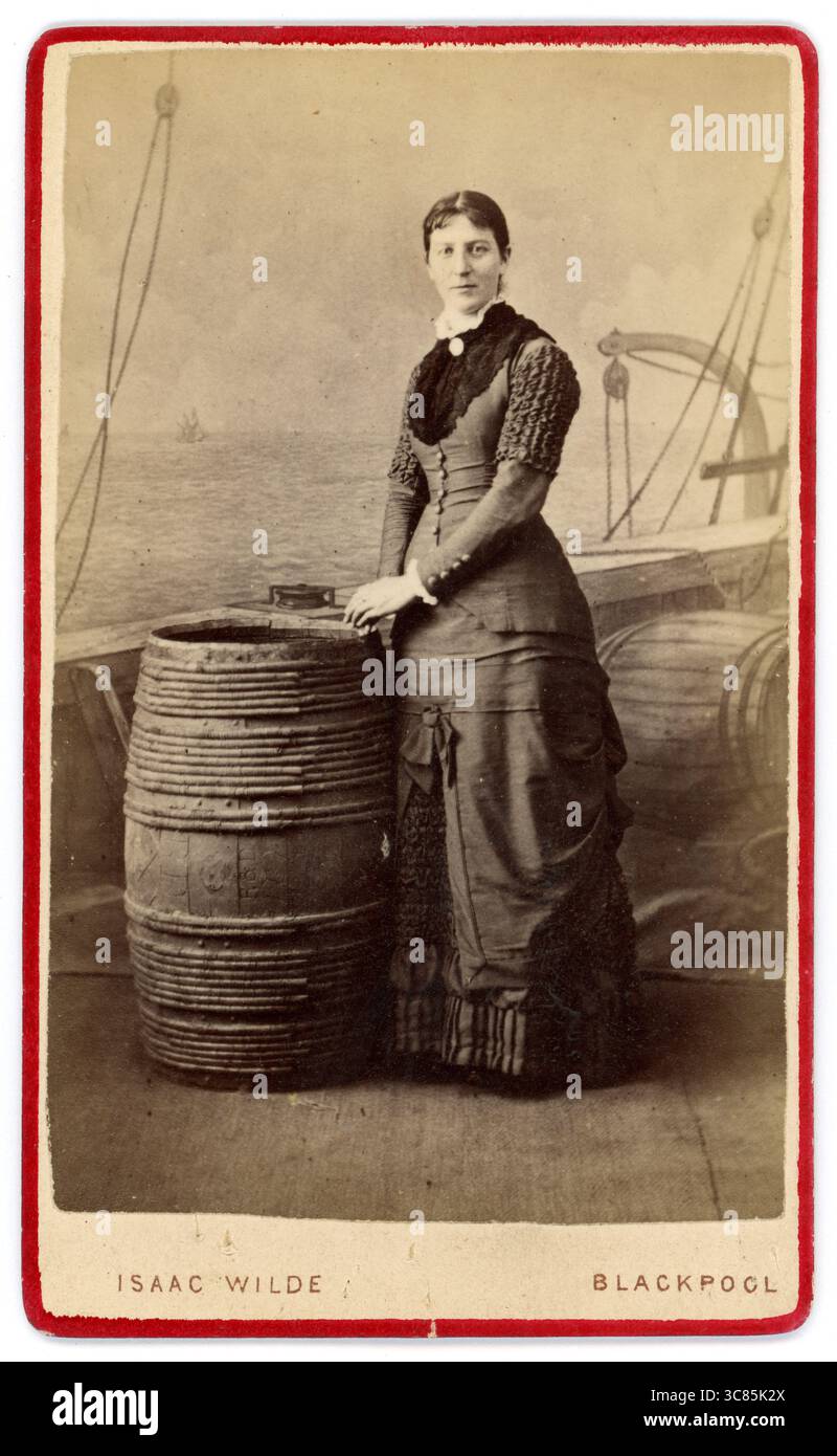 Carta de visite vittoriana originale (biglietto da visita o CDV) di una graziosa donna vittoriana che posa accanto a una botte di quercia, con un ponte della nave e lo sfondo dipinto, è un souvenir per le vacanze al mare dallo studio di Isaac Wilde, 54, Raikes Road, Blackpool, Inghilterra, Regno Unito circa 1880. Foto Stock