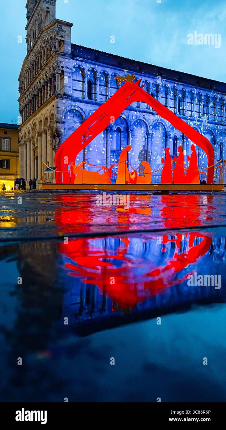 Il presepe natalizio illuminato illumina una piazza della città, riflettendo vividamente sul marciapiede bagnato. Le persone sullo sfondo ammirano lo spettacolo natalizio Foto Stock