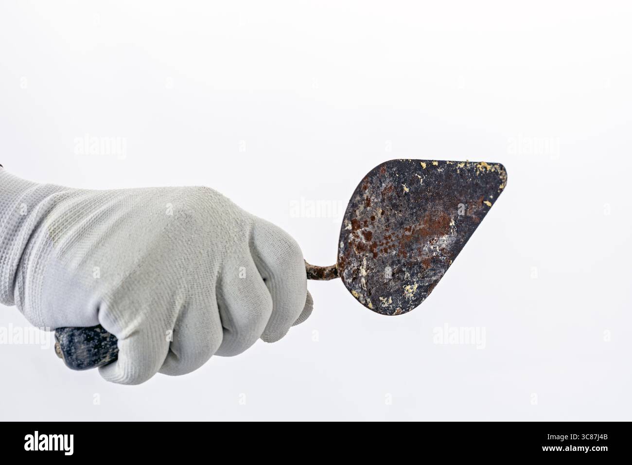 Una mano in un guanto di gomma bianca regge la cazzuola in metallo arrugginito su uno sfondo semplice, suggerendo di concentrarsi sugli utensili da giardinaggio o da costruzione Foto Stock