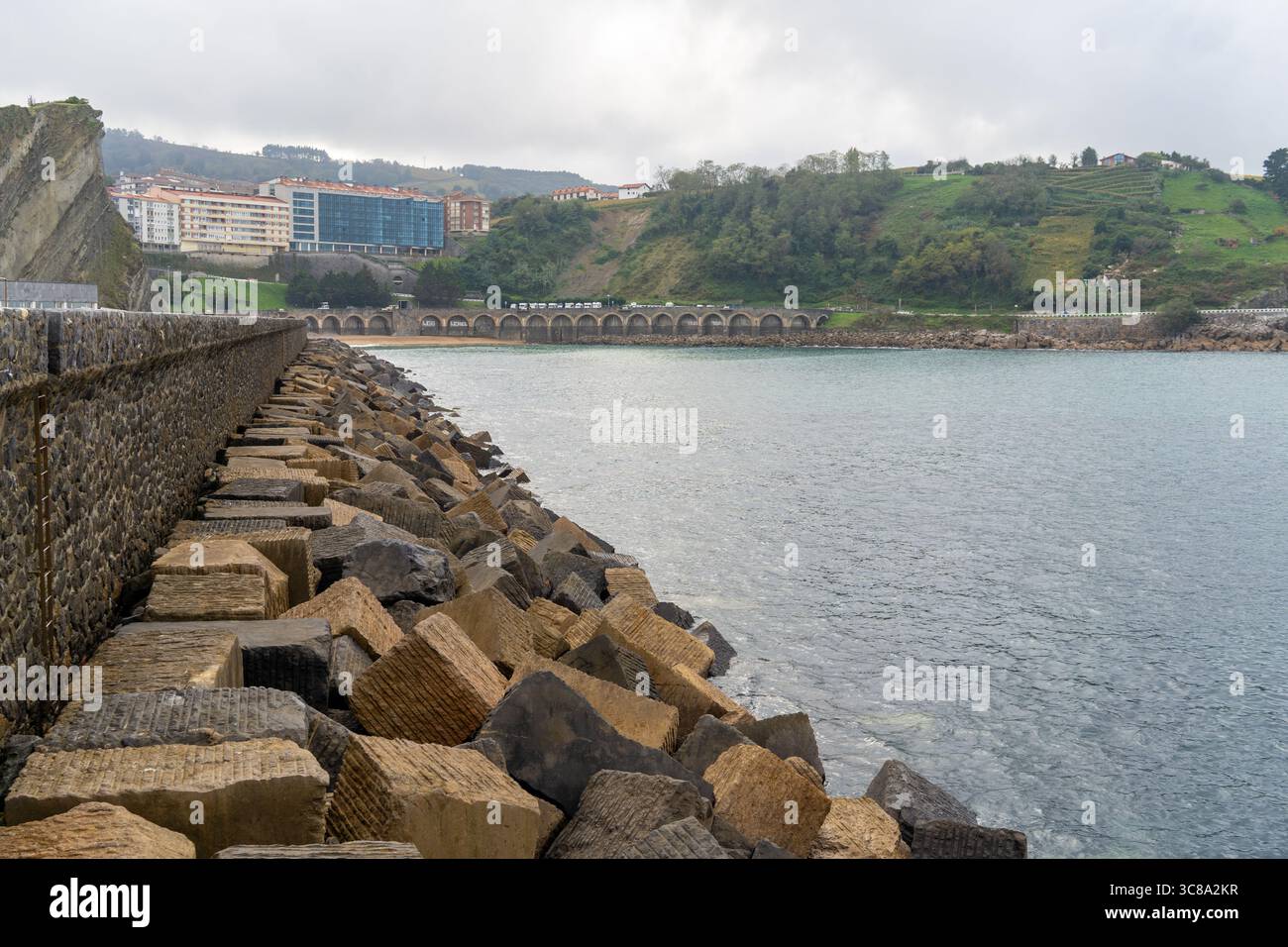 Vista della costa di Zumaia con rocce geometriche e un ponte di pietra distante sotto un cielo nuvoloso, che aggiunge profondità e interesse visivo. Foto Stock