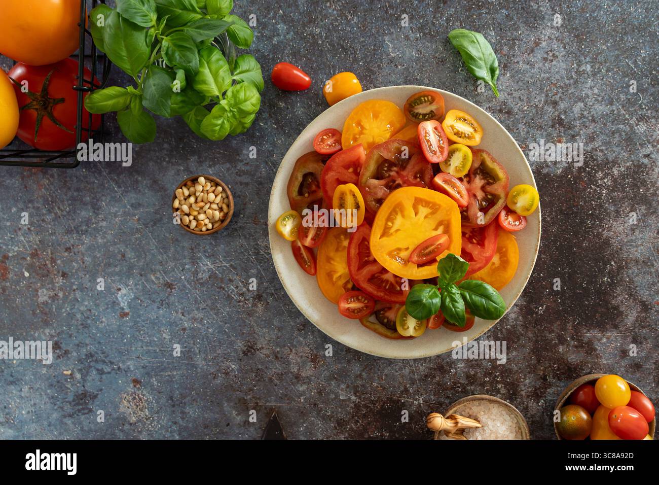 Vista dall'alto dei pomodori colorati a fette con basilico, olive, ciotola di sale e pepe su sfondo marrone blu scuro Foto Stock