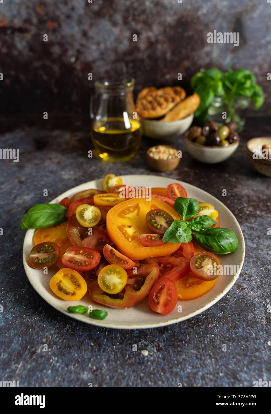 Vista frontale ravvicinata di pomodori assortiti con basilico, bottiglia di olio d'oliva, pane e olive su sfondo rustico scuro Foto Stock