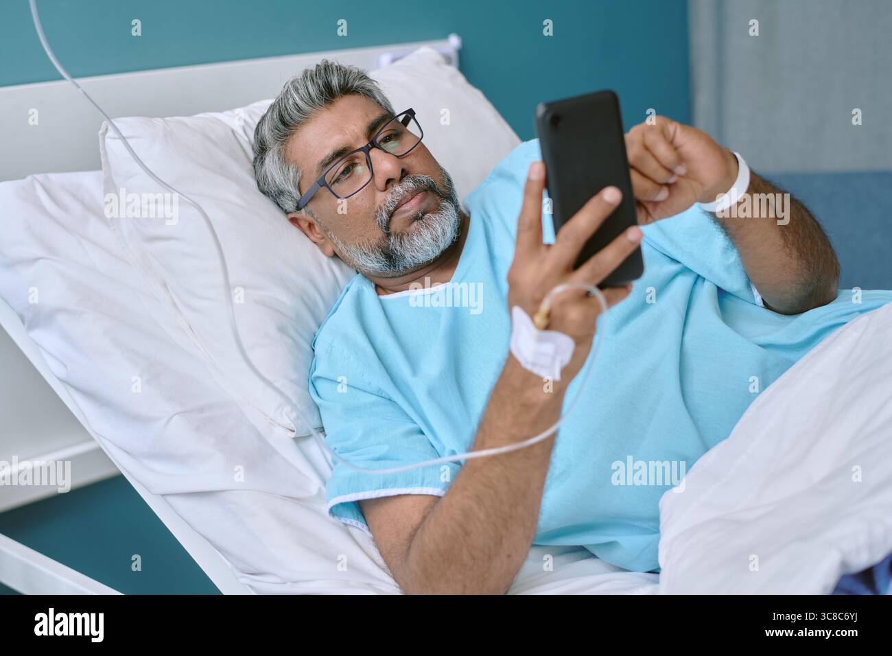 Uomo birazziale di mezza età giacente nel letto di ospedale con dispositivo smartphone Foto Stock