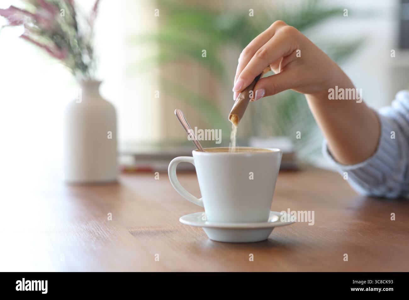 Primo piano di una donna che lancia lo zucchero dal pacchetto in una tazza di caffè su un tavolo di legno a casa Foto Stock