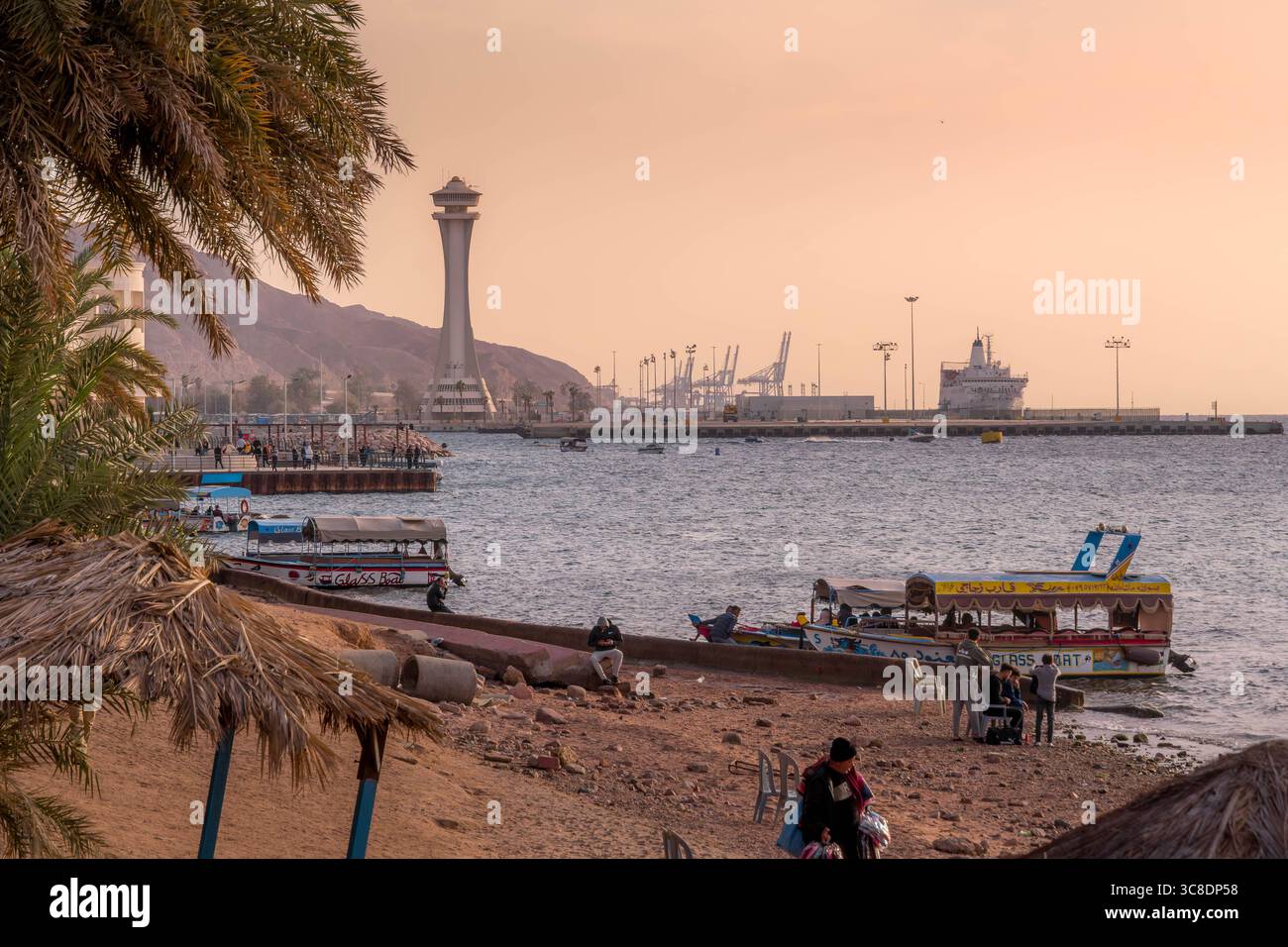 Lungomare di Aqaba e spiaggia con palme, gente del posto, barche in vetro e gru portuali industriali sotto la luce soffusa del tramonto nel sud della Giordania. Foto Stock