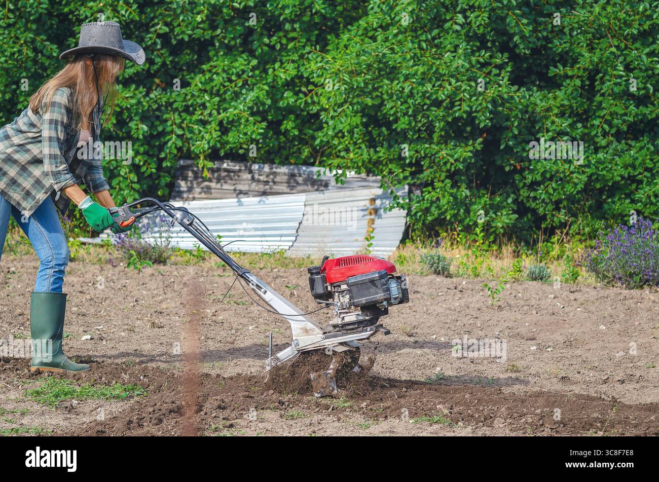 Contadina in cappello e jeans ara il terreno con coltivatore di benzina manuale in giardino, preparando terreni per la semina. Concetto di agricoltura. Foto Stock